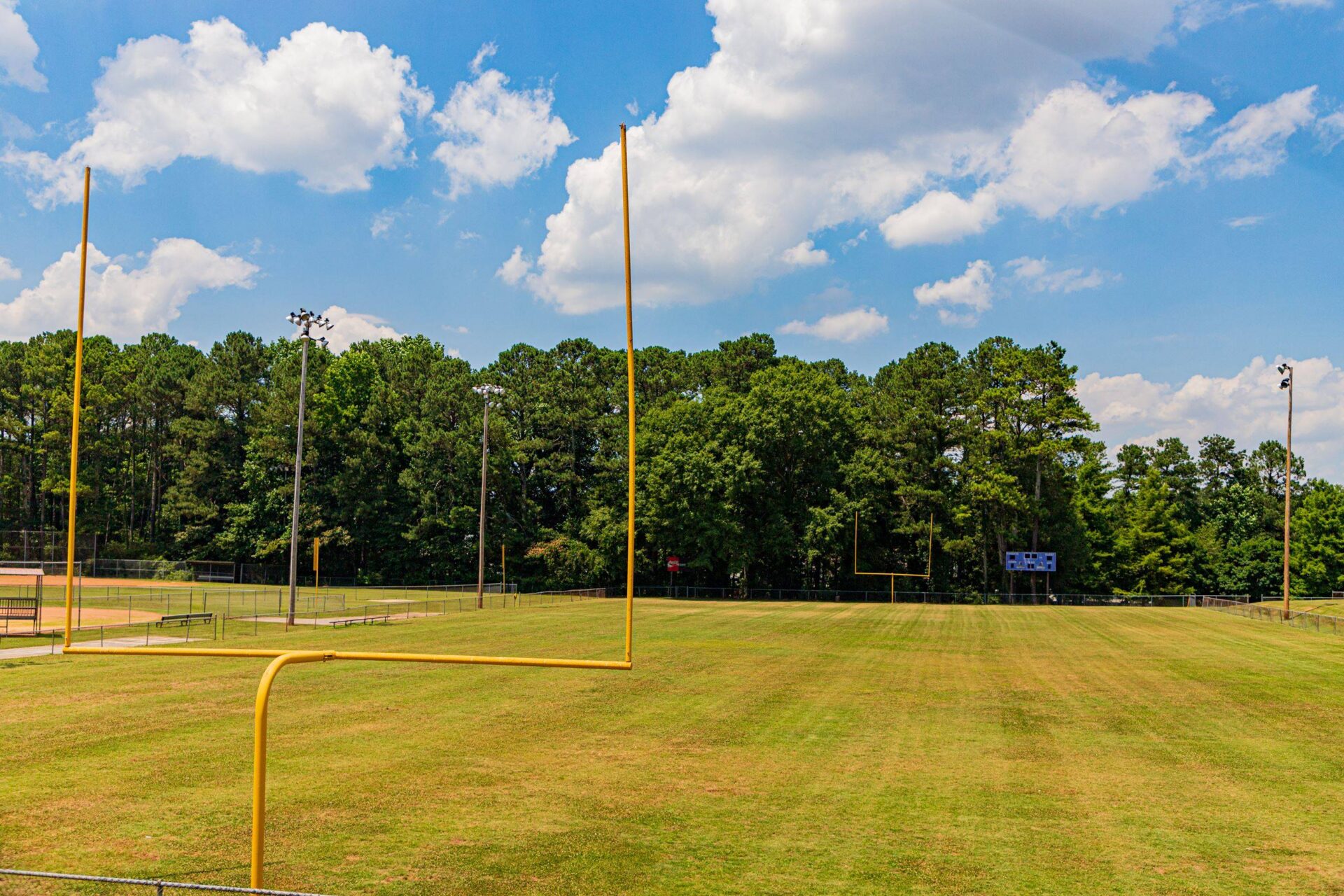 Empty-football-field-with-goalposts-scoreboards-and-lights-surrounded-by-trees-under-a-blue-sky