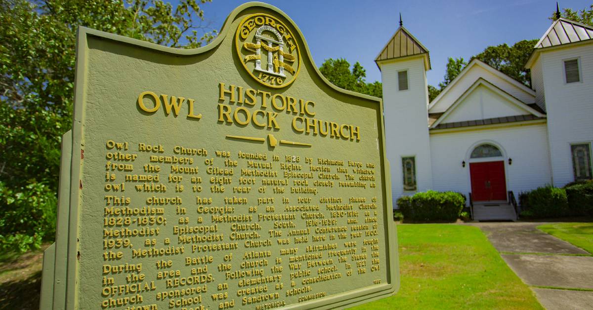 Historical marker for _Owl Rock Church_ in front of a white church with a red door under a blue sky.