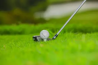 A golf ball teed up on lush green grass, ready to be struck by a silver club in the background.